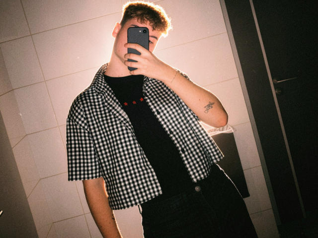 MarcusIg69 with a checkered shirt, black tee, and dark pants, taking a selfie with a phone in a dimly lit bathroom, showing a tattoo on his right arm.