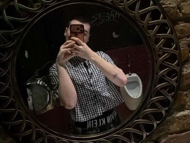 MarcusIg69 with glasses, checking mirror selfie, holding phone, wearing black-and-white checkered shirt, tattoo on right arm, reflection in ornate mirror, urinal in background.