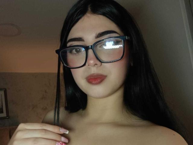 Isabella Rossi, con lunghi capelli neri, che indossa grandi occhiali neri e rossetto rosso, ha un'espressione leggermente corrucciata ed è senza top, con una mano vicino alla clavicola..