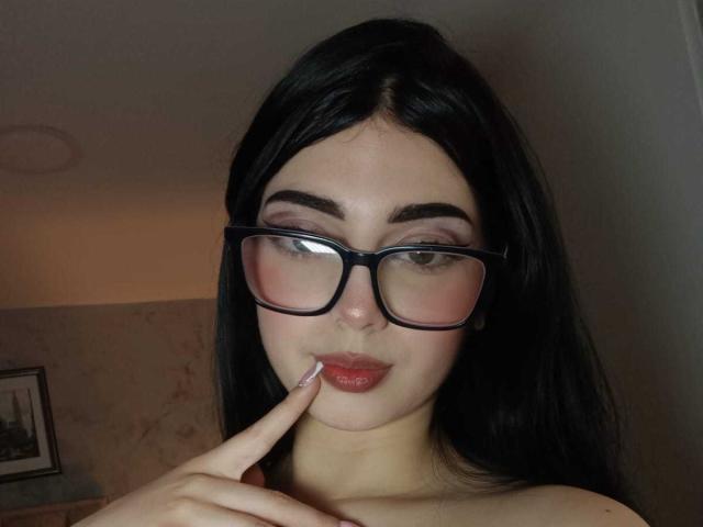 Isabella Rossi, avec des lunettes noires, des cheveux foncés, un fard à paupières rose et un rouge à lèvres rouge, touche ses lèvres rosâtres avec son doigt. Elle a une apparence mignonne, légèrement studieuse..