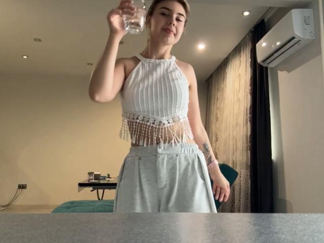 SlyviaCoffen with a white crochet top, fringe, and grey pants, holding a water bottle, her tits visible, standing in a room with a green chair and air conditioner.