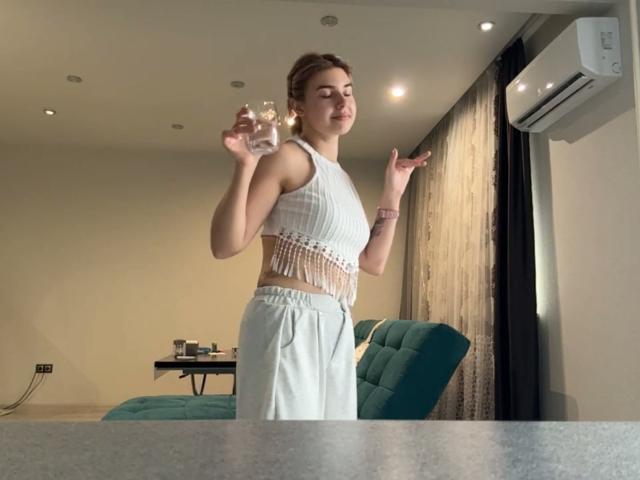 SlyviaCoffen with light brown hair in a white fringe crop top, holding a glass, wearing white pants, standing in a room, air conditioner above, green chair behind.