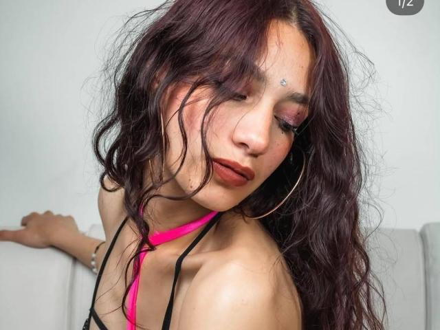 AmberVegas mit welligem, lila Haar, einem pinken Halstopp, Reifenohrringen und rotem Lippenstift, die leicht grinsend nach unten schaut. Sie hat ein kleines Tattoo unter ihrem rechten Auge..