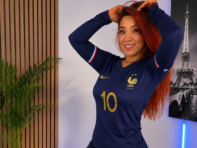 JannaLorenstmd, mit roten Haaren, lächelnd, trägt ein dunkelblaues Fußballtrikot mit der Aufschrift "10" und der französischen Flagge, hat die Hände im Haar, steht, zeigt ihre Brüste leicht, in einem Raum..