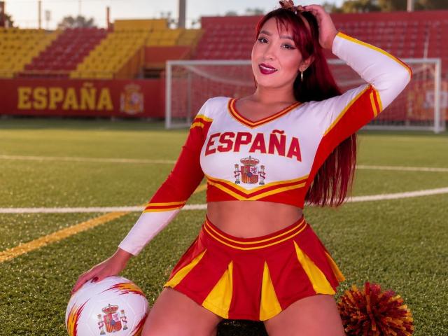 JannaLorenstmd con un completo da cheerleader rosso e giallo, che mostra il seno, che tiene un pallone da calcio, seduta sull'erba, con una mano tra i capelli, sorridente, con lunghi capelli rossi, rossetto rosa..