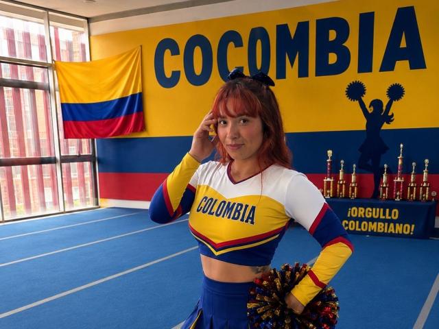 JannaLorenstmd ha i capelli rossi, indossa un'uniforme da cheerleader colombiana con i colori giallo, blu e bianco, tiene in mano dei nastrini, è in piedi su un tappetino blu, con una bandiera e dei trofei sullo sfondo..