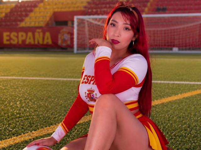 JannaLorenstmd con i capelli rossi, seduta su un campo da calcio, indossa un completo da cheerleader spagnola rosso e giallo, mostrando il seno, con una mano vicino alla bocca..