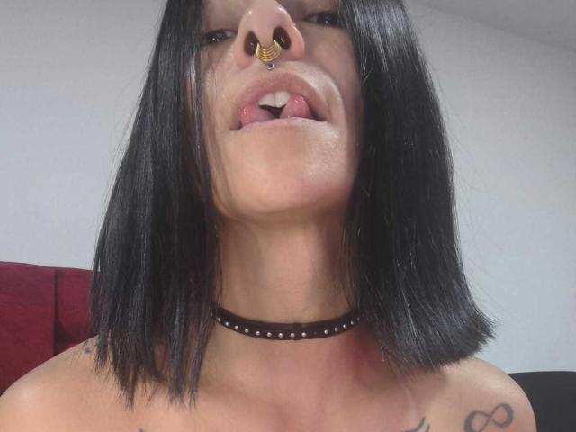 Amelie Blake, con i capelli neri a metà schiena, un piercing al naso, la lingua fuori, un collare nero e il petto tatuato, che mostra i suoi seni e il piercing al naso..