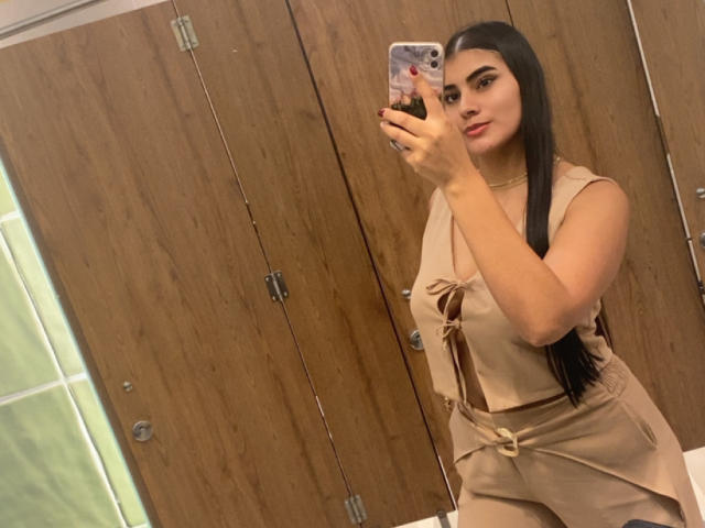 AlaraKey with long black hair, wearing a beige dress with brown straps, taking a selfie in a mirror, holding a phone with a floral case, showing her ass.