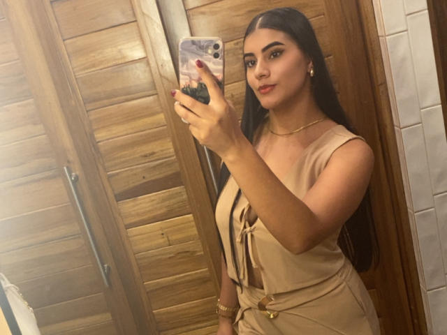 AlaraKey with long black hair, tan dress, and gold necklace, holding phone, taking mirror selfie, showing her tits and ass in warm wooden bathroom.
