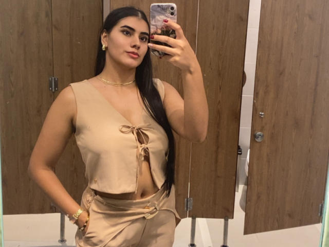 AlaraKey with long black hair, tan blouse with tied front, and tan pants, taking a mirror selfie in a bathroom, wearing gold earrings and a necklace. Boobs visible through blouse.