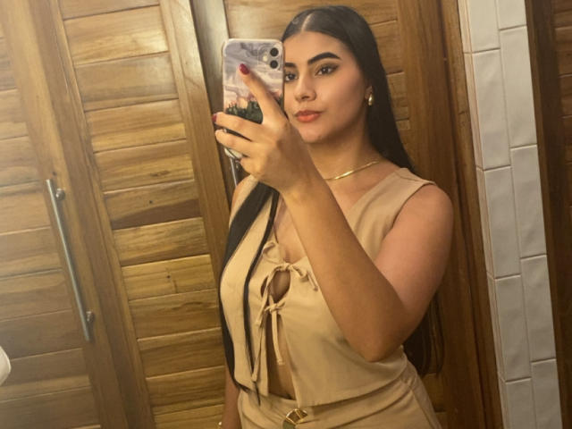 AlaraKey with long black hair, beige dress with keyhole, showing tits, holding phone in mirror, wood shutters, gold choker, red nails, smiling, brown eyes, in selfie.
