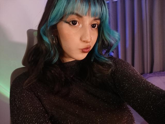 CatBleu, avec ses cheveux ondulés bleu turquoise et noirs, porte un haut noir scintillant à manches longues, a un anneau au nez et un eye-liner ailé, et regarde l'appareil photo avec une expression boudeuse..