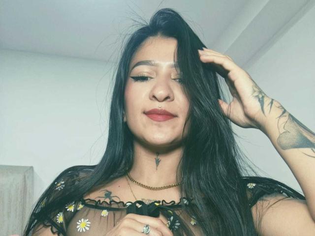 LuciadelaVega with long black hair, wearing a sheer black top with daisy patterns, touching her hair, tattooed arm, gold necklace, red lips, slight smile, and a ring on her finger.