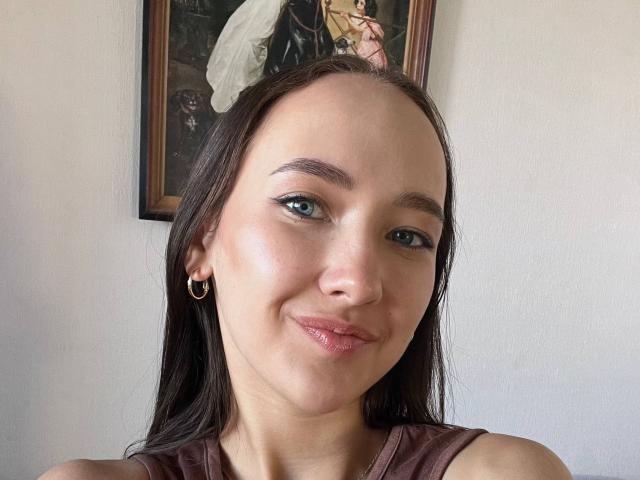 Valorie Santulli, mit blauen Augen, braunen Haaren und einem leichten Lächeln, trägt ein braunes Top ohne Ärmel, kleine goldene Ohrringe und einen kleinen Nasenring. Ihr Gesäß ist nicht sichtbar..