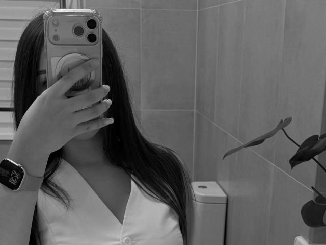 Tawnya Kaczmarski, avec de longs cheveux foncés, tenant un téléphone, se cachant le visage, portant une chemise blanche et une montre intelligente noire, dans une salle de bain. Le reflet montre des toilettes et une plante en pot. Image en noir et blanc..