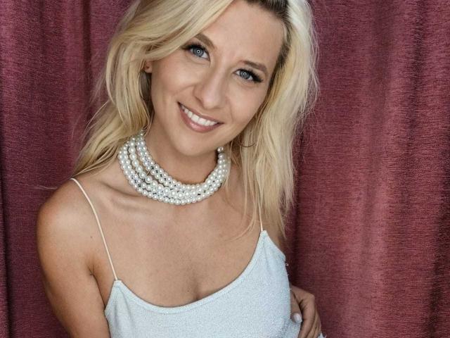 KaylinJan with blonde hair, blue eyes, and a smile, wearing a sparkly white dress and multiple pearl necklaces, showing her small tits.