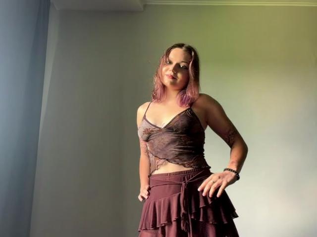 LeanaBairos with pink-dyed hair, wearing a sheer brown top and ruffled maroon skirt, standing in a room with sunlight, hands on hips, smiling confidently.