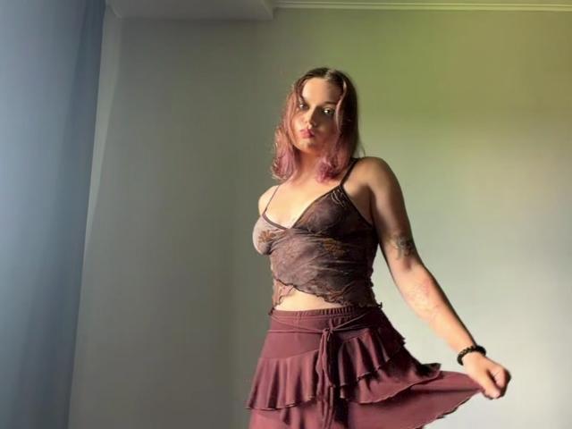 LeanaBairos with pink-dyed hair, wearing a sheer brown lace top and a ruffled maroon skirt, standing in a room, showing off her small tits and tattooed arm.