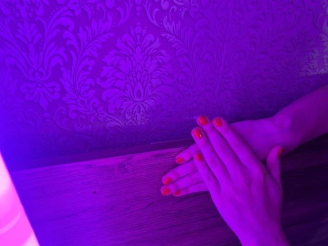 WenTina's hands with orange nails are resting on a wooden surface, bathed in purple light, showing her delicate fingers and slightly curled pinky.