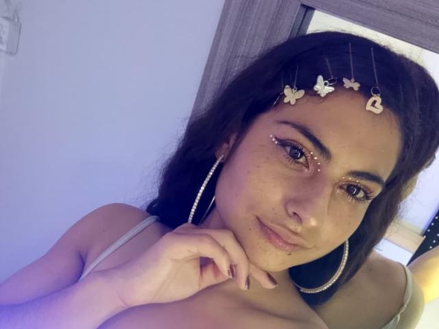 AngelAlba with brown hair, wearing a headpiece with butterflies and hearts, has freckles, eyeliner, and a nose ring, smiling with hoop earrings, and resting her chin on her hand.