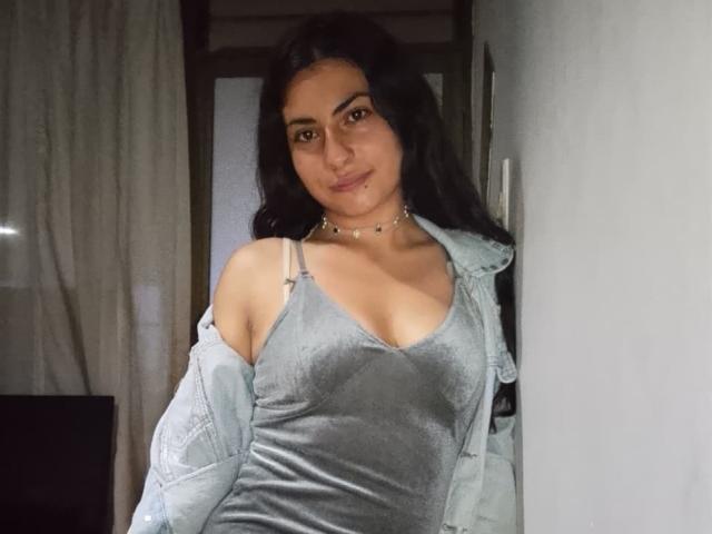 AngelAlba with long black hair, wearing a gray velvet dress, light denim jacket off her shoulders, showing her small tits, and a subtle smile, standing near a white wall.