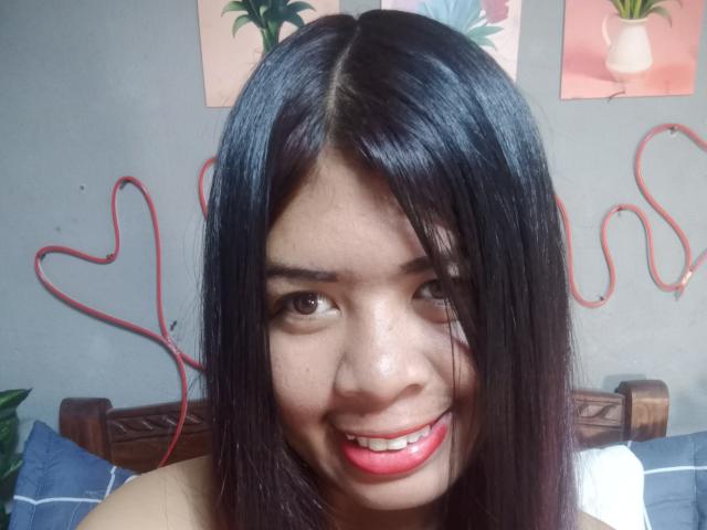 MariahSquirting69 ha i capelli neri e lisci, gli occhi marroni e un sorriso rosa con rossetto ben visibile. Un cordoncino rosso a forma di cuore le pende dietro la testa. Sta sorridendo alla fotocamera..