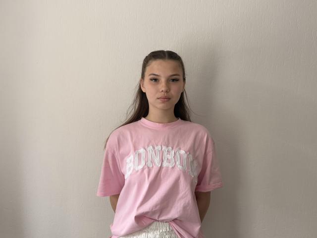 Clementina Swantek, avec de longs cheveux bruns, un t-shirt rose avec l'inscription "BONNET" et un pantalon blanc, se tient devant un mur uni, regardant droit devant la caméra, les mains dans les poches..