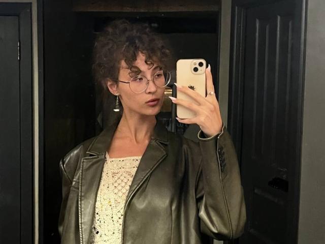 AlyceErway, con i capelli castano ricci, gli occhiali e una giacca di pelle verde, tiene in mano un telefono, indossa una camicetta bianca in pizzo e sta scattando un selfie allo specchio, guardando verso la fotocamera..