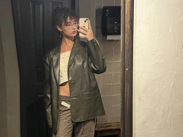 AlyceErway, com cabelos castanhos cacheados, óculos e um casaco de couro sobre uma blusa cropped branca de renda, segurando um telefone em um selfie no espelho, mostrando uma silhueta magra e discreta..