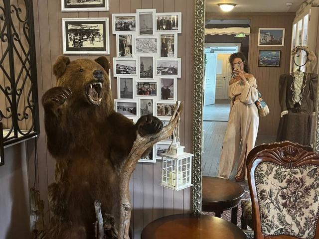 AlyceErway prend un selfie devant un miroir, debout à côté d'une statue réaliste d'un ours brun, dont la bouche est ouverte. Elle porte une combinaison beige et des talons rouges..