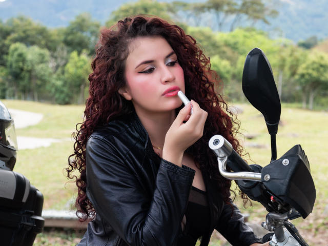Thaliaa, uma mulher com cabelo ruivo cacheado, vestindo uma jaqueta de couro preta, aplicando batom rosa em seus lábios cheios, sentada em uma motocicleta com um espelho em primeiro plano..