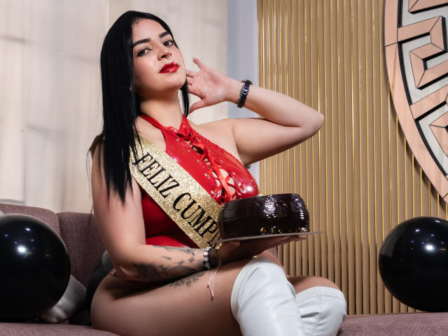 LilithAmoth met zwart haar, een rode haltertop, witte laarzen, een lint, die een chocoladetaart vasthoudt, een getatoeëerde arm, zittend op een bank, zwarte ballonnen, een zelfverzekerde glimlach, rode lippenstift, de linkerhand die de wang aanraakt..