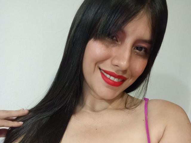 MadissonFontaine has long black hair, red lipstick, and pink eye shadow, smiling with visible teeth, wearing a pink strap, touching her hair with her right hand.