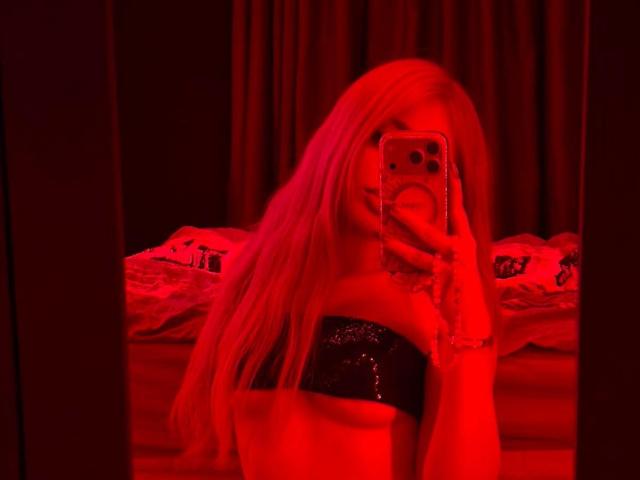 Selfie von AvaCharm mit rotem Licht, mit langen blonden Haaren, die ein schwarzes, glänzendes Top ohne Träger trägt, wobei sie ihr Gesicht mit einem Handy verdeckt, Tätowierung am Handgelenk, Spiegelbild..