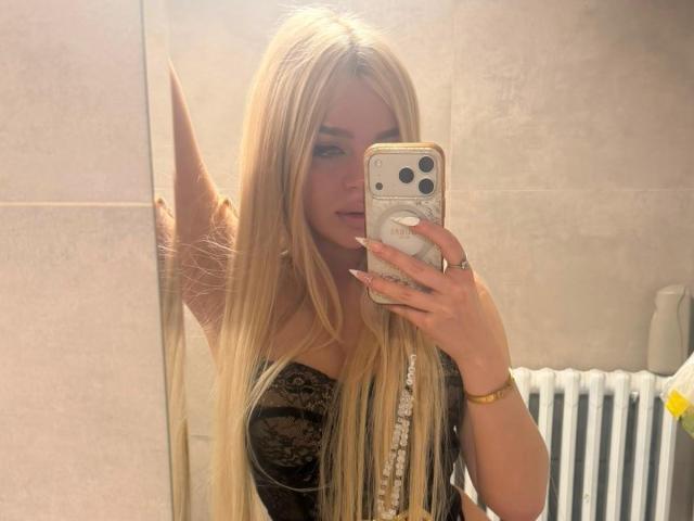 AvaCharm, con i suoi lunghi capelli biondi, una maglia di pizzo e un anello, sta scattando un selfie allo specchio con il suo telefono, mostrando il seno e un gluteo leggermente visibile in un bagno..