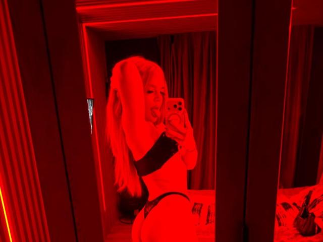 AvaCharm macht ein Selfie vor einem spiegelnden roten Licht, wobei ihr Hintern in schwarzem String-Unterwäsche zu sehen ist. Sie hat langes blondes Haar, lächelt und hält ein Handy, im Hintergrund ist ein schwach beleuchteter Raum zu sehen..