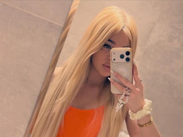 AvaCharm mit langen blonden Haaren, einem orangefarbenen Oberteil, die ein Telefon vor einem Spiegel hält und ihr Gesicht und ihre Hand mit einem Haargummi zeigt, blickt in die Kamera..