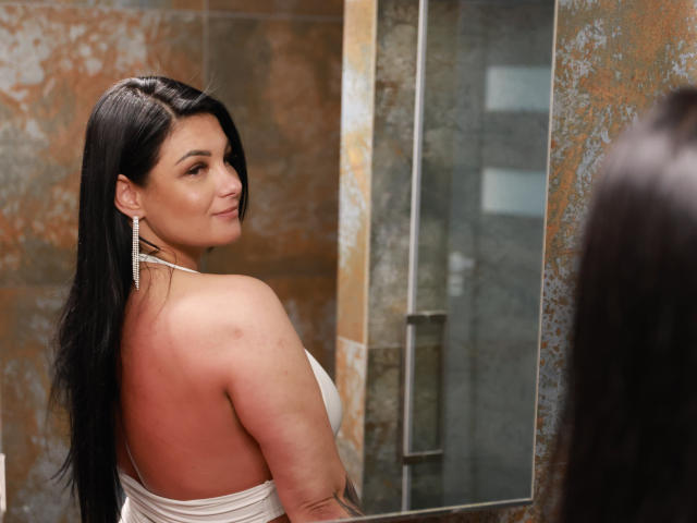 IvyMidnight with long black hair, white strapless dress, and earrings, looking back at a mirror, showing her tits and a small tattoo on her right arm.