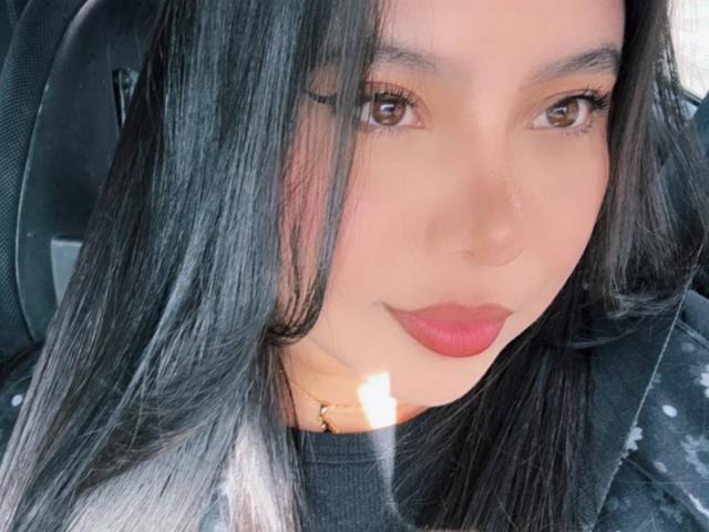 NoaBlossoms Gesicht, braune Augen, langes schwarzes Haar, rote Lippen, trägt ein schwarzes Oberteil, Sonnenlicht auf der rechten Wange, leichtes Lächeln, sitzt, wobei ein schwarzer Autositz im Hintergrund sichtbar ist..