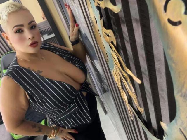 Gianella Cox, com cabelo loiro platinado, decote profundo em uma blusa listrada, tatuagem no ombro direito, unhas vermelhas, pulseiras douradas, em pé em frente a uma parede coberta de grafites..