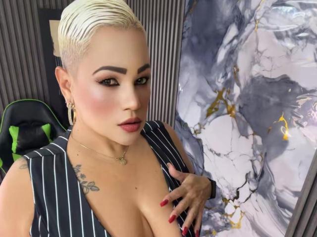 GianellaCox, avec des cheveux blonds courts, un maquillage foncé, du rouge à lèvres rouge, un haut à rayures fines, une main sur ses seins, un tatouage sur la poitrine gauche, des boucles d'oreilles dorées, des ongles rouges, assise sur une chaise..