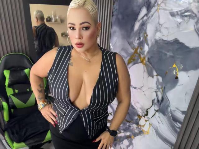 GianellaCox has big tits, wearing a black and white pinstriped top, blonde hair, tattoos, red lipstick, and a black chair with green accents in the background.