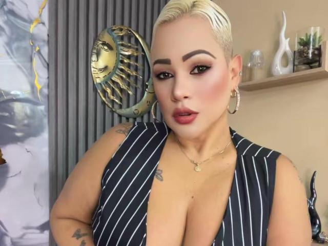 Gianella Cox, con i capelli biondi corti, un seno abbondante, indossa una camicetta a righe, orecchini a cerchio e una collana, ha un tatuaggio sul braccio destro e un'espressione accigliata..