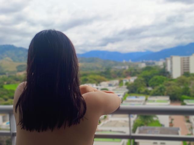 Topless Genesiss with shoulder-length dark brown hair, arms crossed, facing a city view with mountains and cloudy sky in the background. Her back is to the camera.