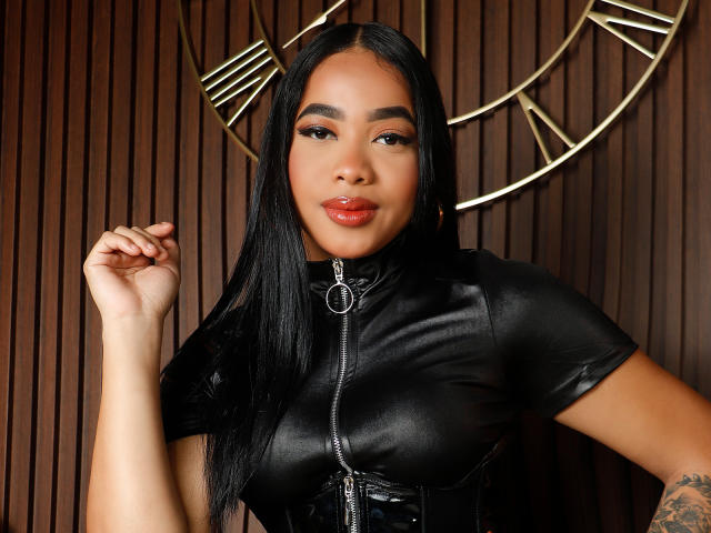 KarlaBella, con cabello negro largo, vestida con una blusa negra de cuero con cremallera, que deja ver sus senos, y un tatuaje en su brazo derecho, tiene labios rojos y una expresión segura..
