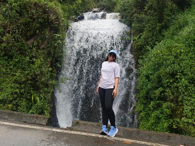 ZaraBlaze stands in front of a waterfall, wearing a white tee, black pants, blue sneakers, and a blue cap, with greenery on both sides and a wet road below.