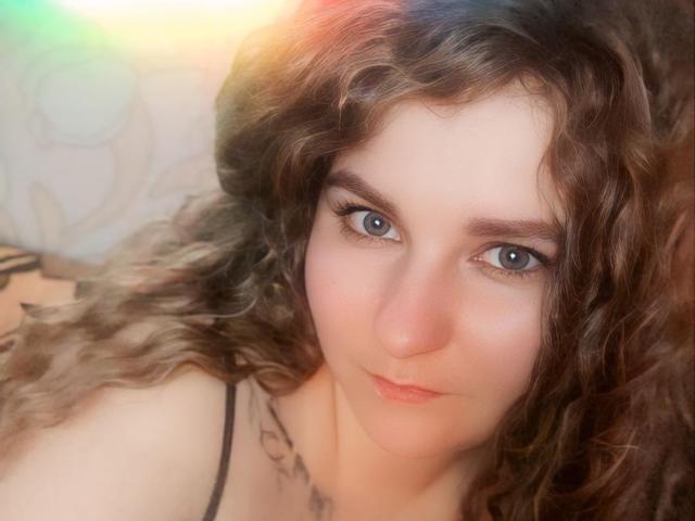 MinistraX has curly brown hair, blue eyes, and a light skin tone, with subtle makeup and a slight smile. She's wearing a black spaghetti strap.