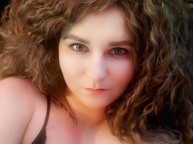 MinistraX has curly brown hair, pale skin, and blue eyes. She's wearing a black spaghetti strap top, with a tattoo on her left shoulder. Her boobs are partially visible.