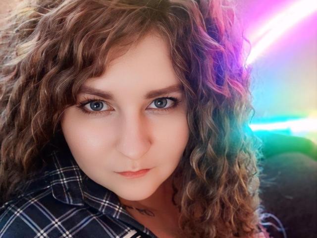MinistraX has curly brown hair, blue eyes, and a neutral expression. She's wearing a plaid shirt with a subtle tattoo on her neck. Bright neon light in background.